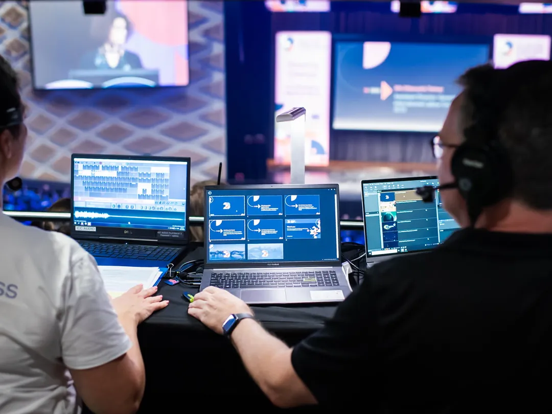 AV technician with a headset at a laptop control desk, watching the session feed