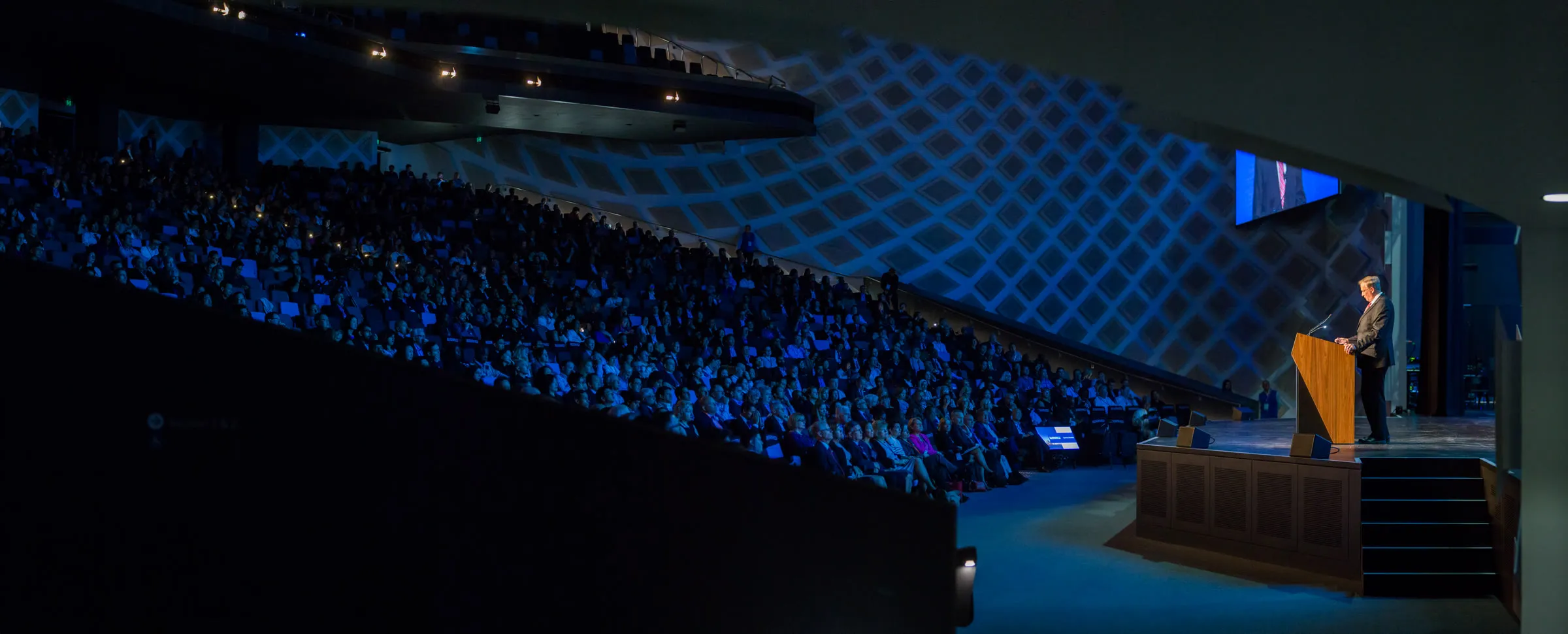 Ultra-wide view of a dark plenary theatre, a speaker at the lectern in front of a full audience